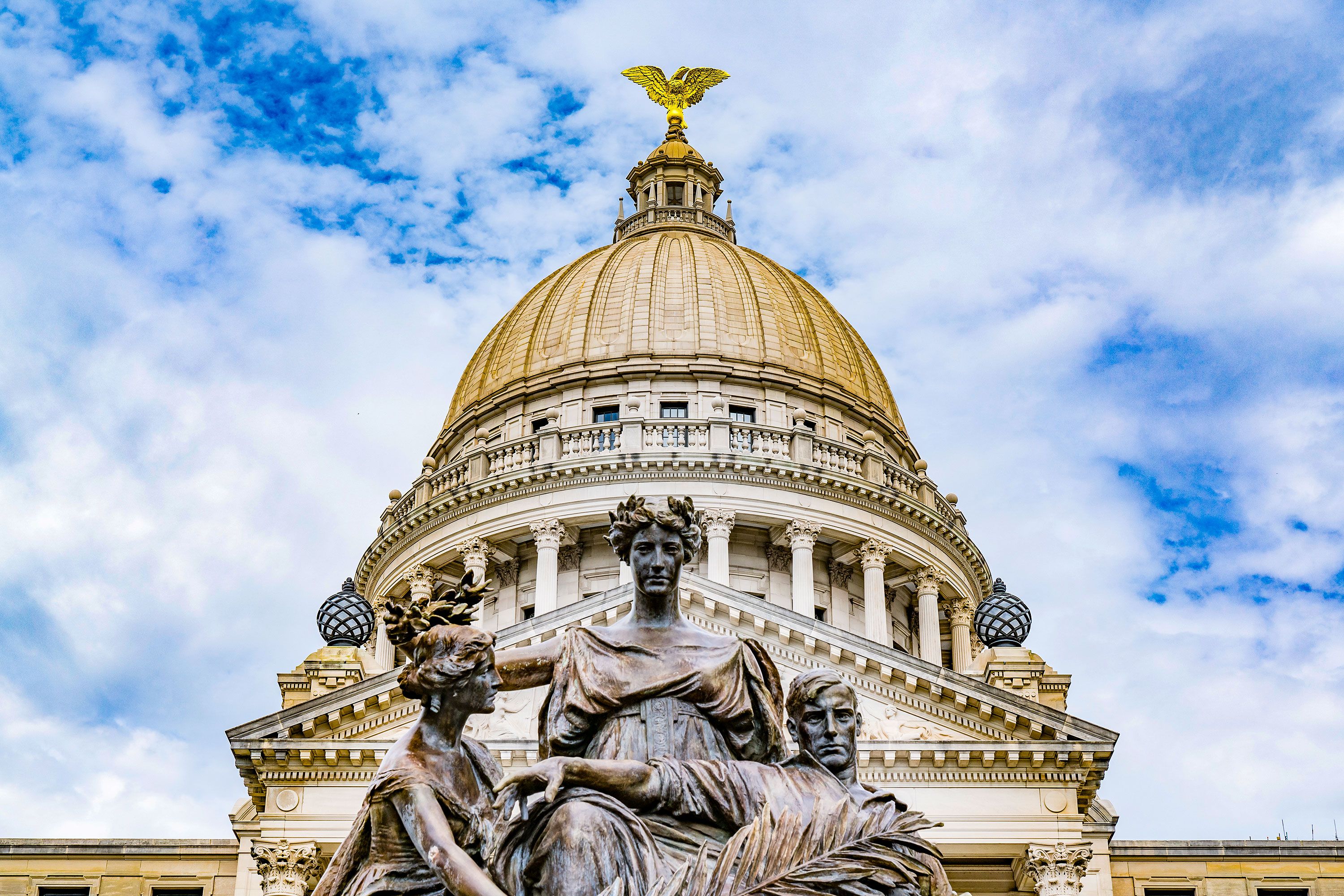 A three-person statue with laurels in front of a domed government building with a golden eagle at the top.