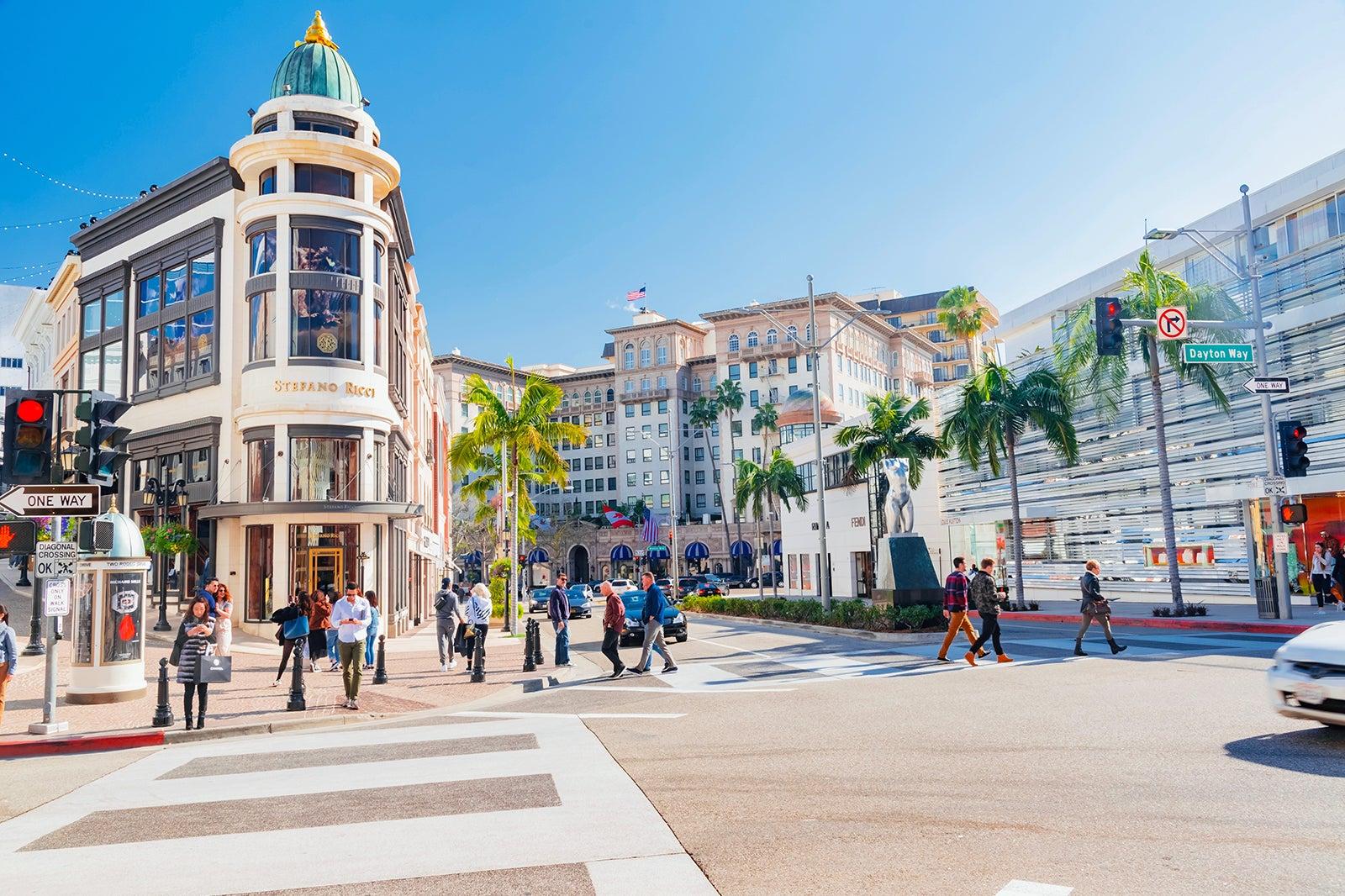An interstection in LA with tall buildings, palm trees, and people using the crosswalks.