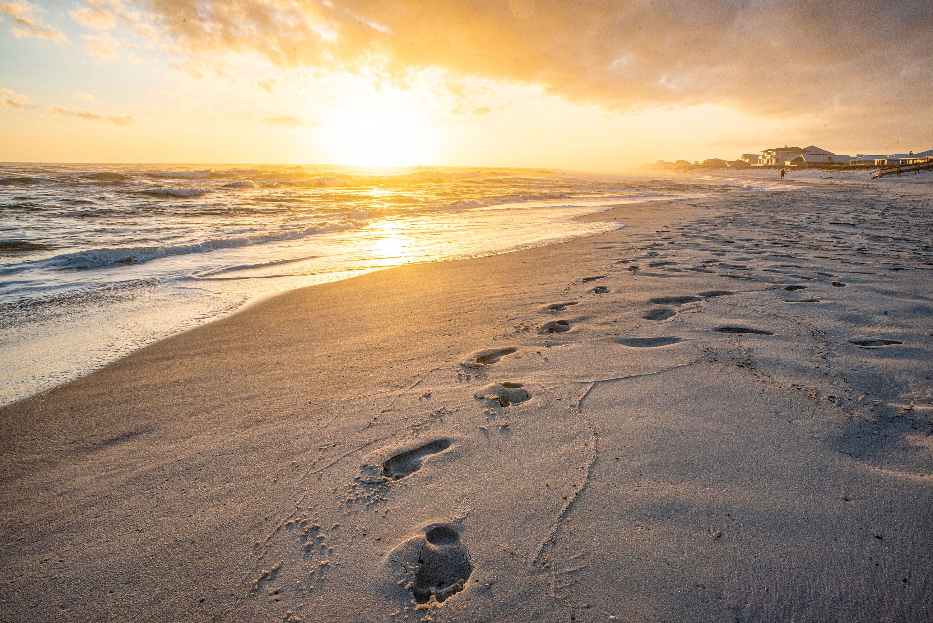 Footprints in the sand walking towards frame on a beach with gentle waves and a building in the distance at sunset.