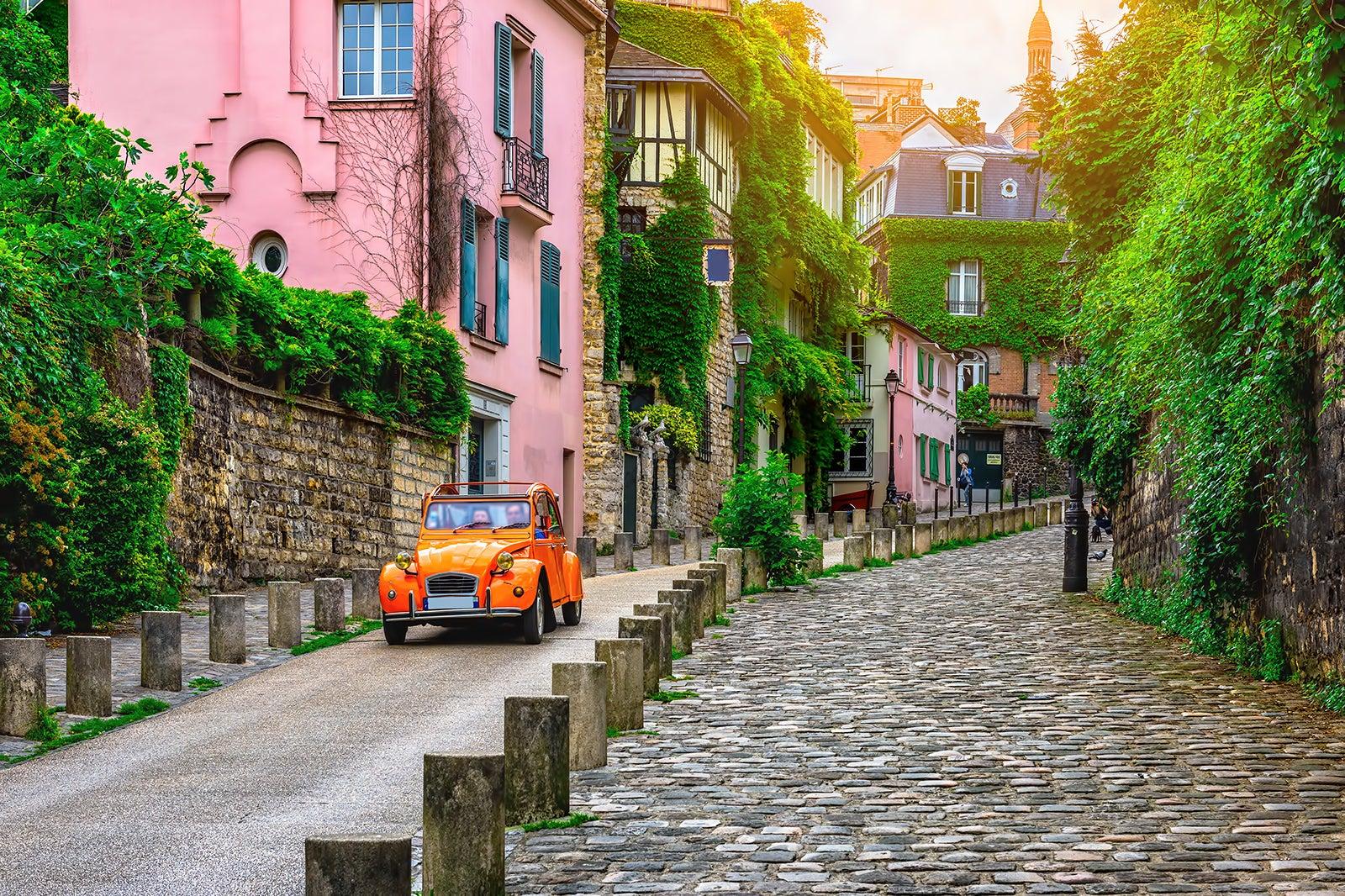 Small orange car on one-lane street next to cobblestone walkway with pink homes and ivy on brick wall.