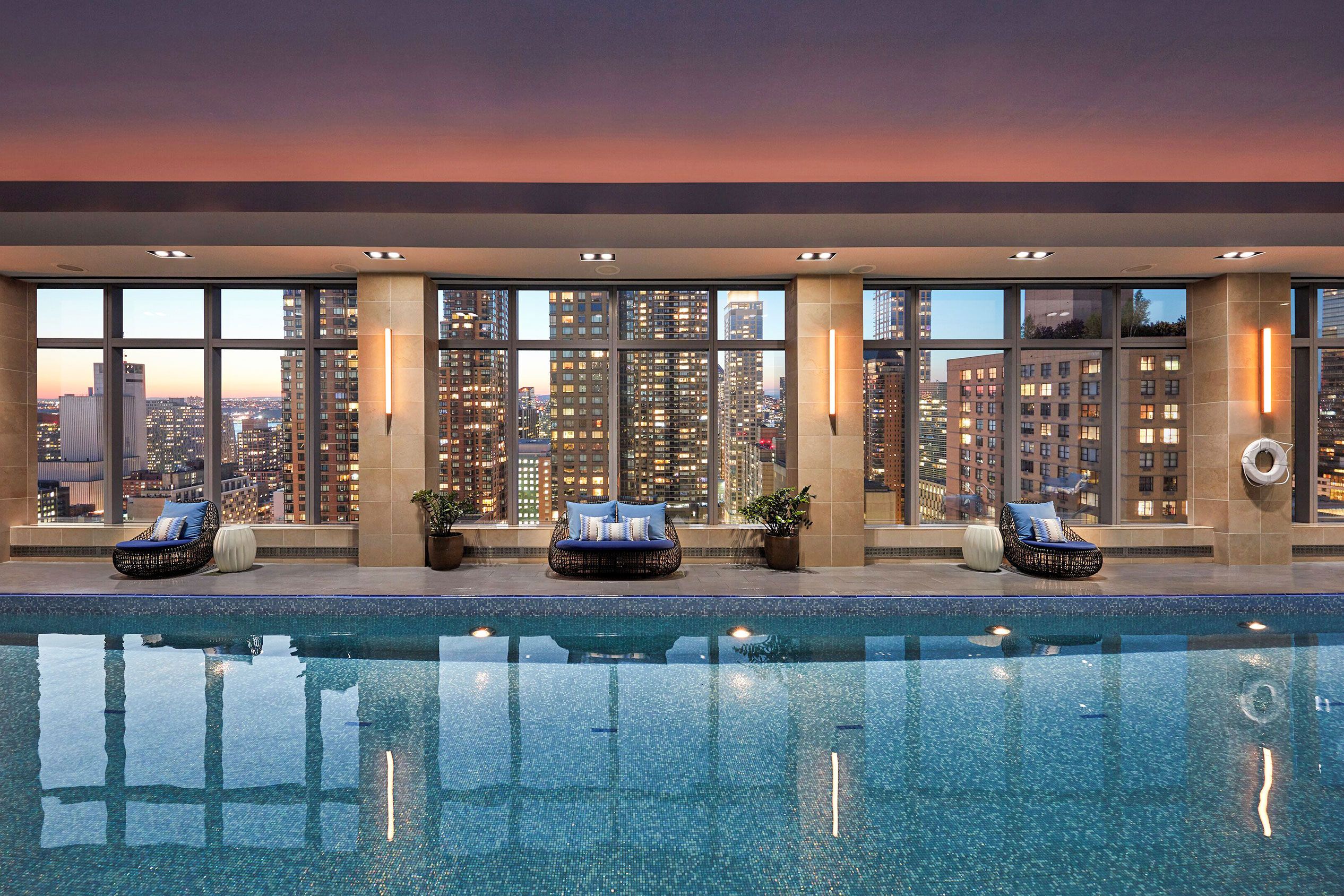 An indoor pool with floor-to-ceiling windows and a view of the New York City skyline.