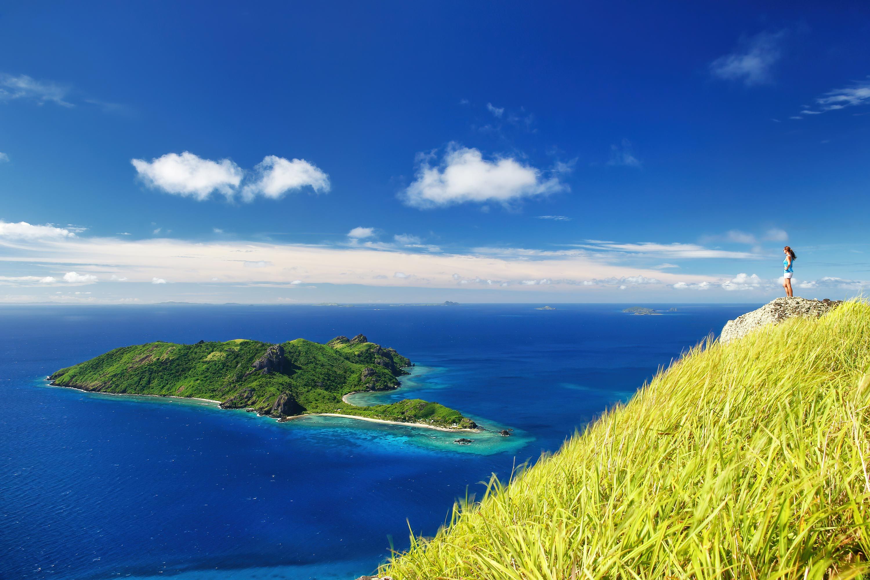 View of a woman standing on a rock surrounded by bright lime green grass looking out over the ocean and an island in the distance in Fiji.