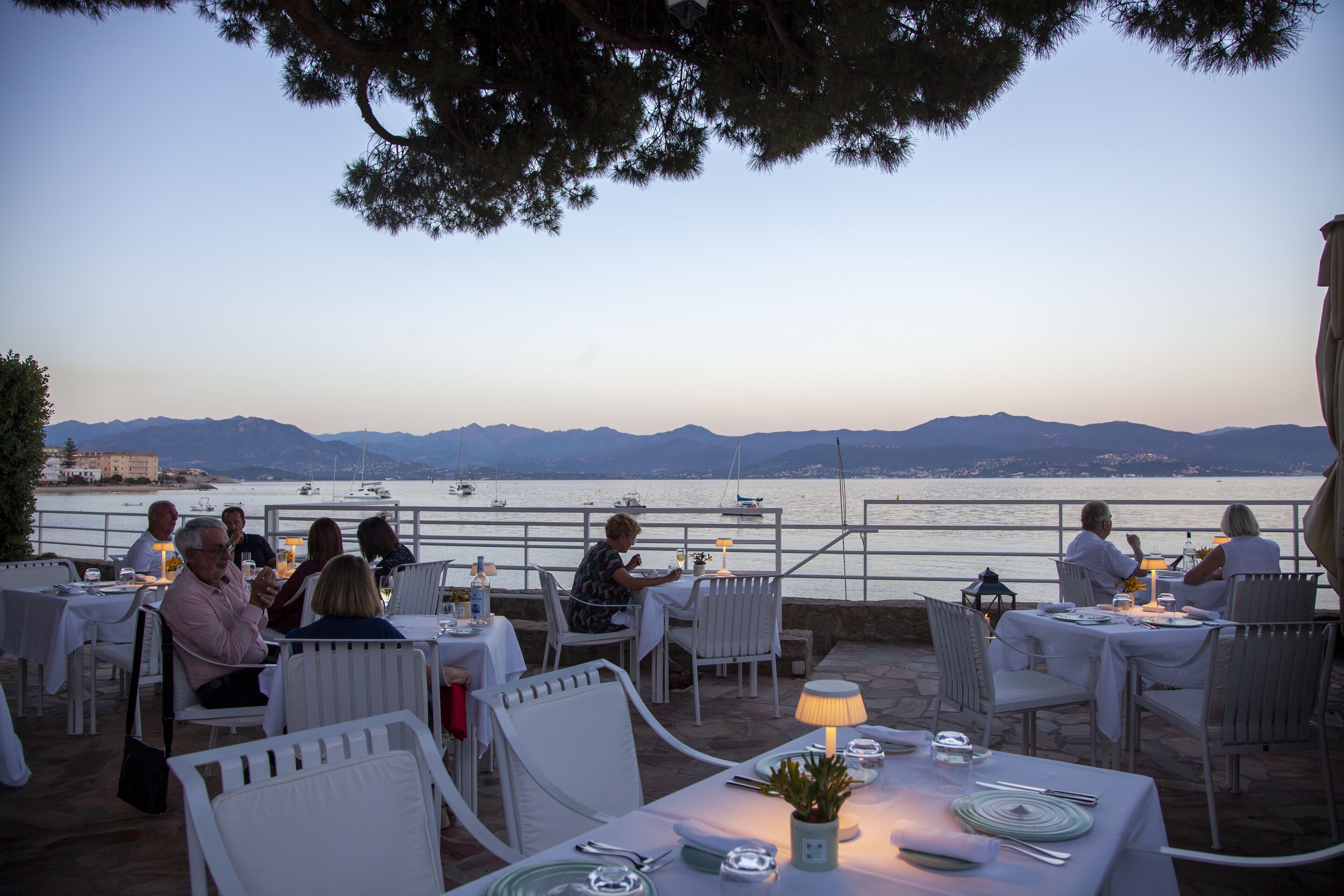 People sit at white tables with a coastal view at a restaurant.