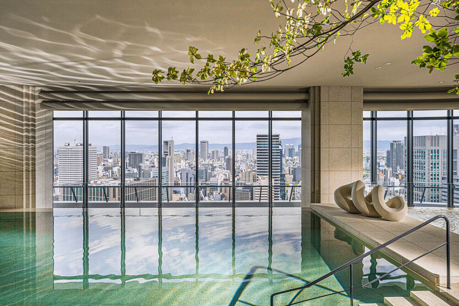 A sea green indoor pool with ceiling-to-floor windows overlooking a city skyline view. 