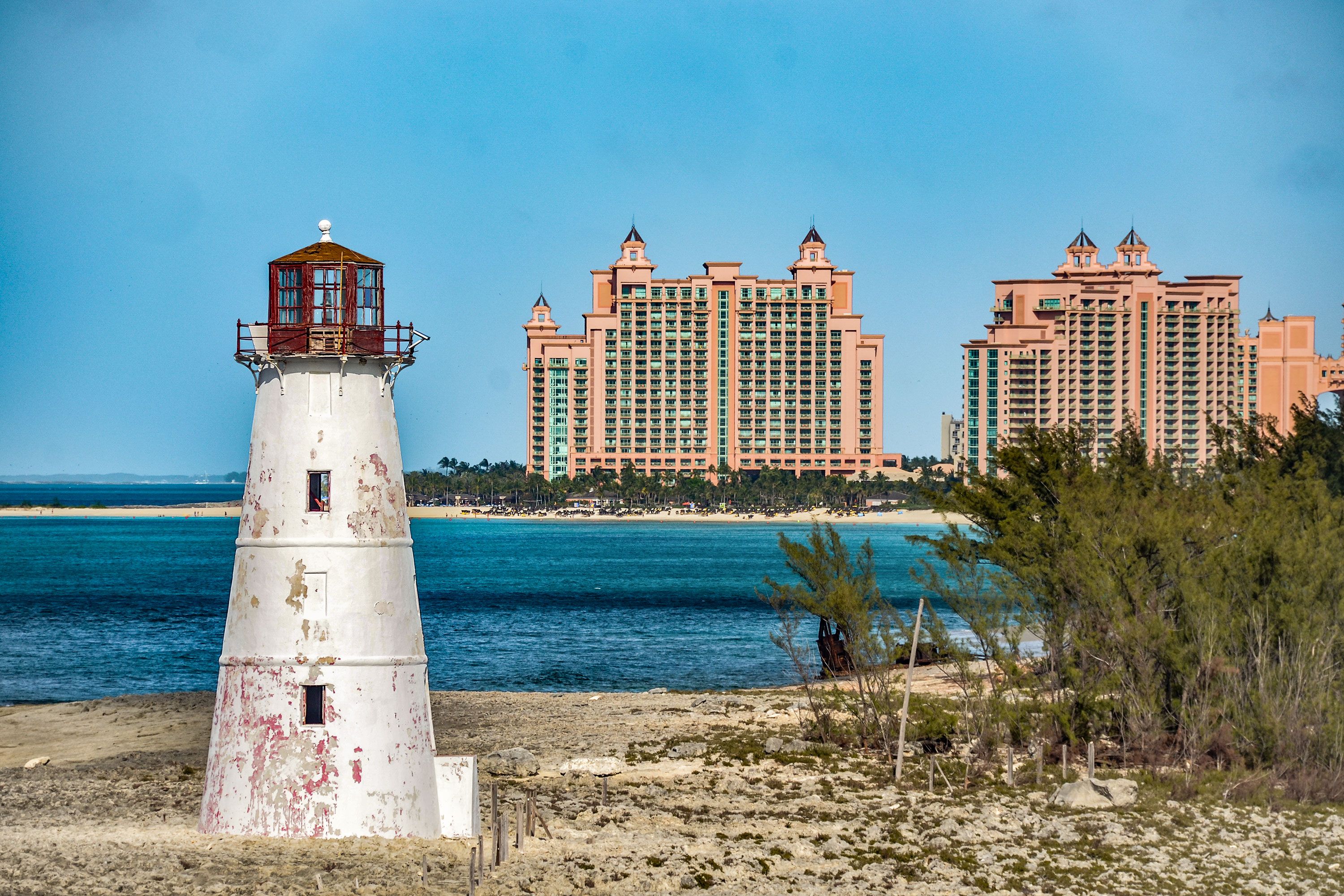 Weathered lighthouse with two large resort buildings in the distance past blue water and beach.
