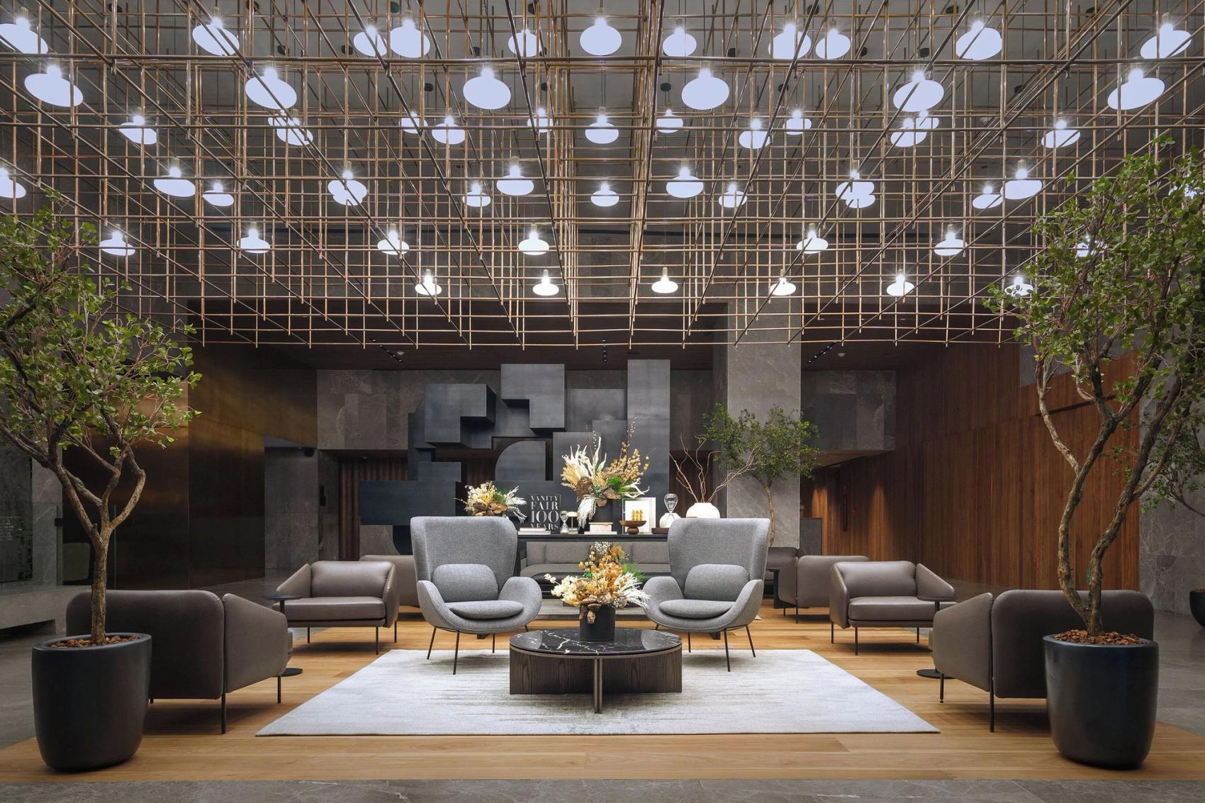 A lounge area with modern furnishings, potted trees, elaborate metal ceiling feature and grey stone floor and walls.