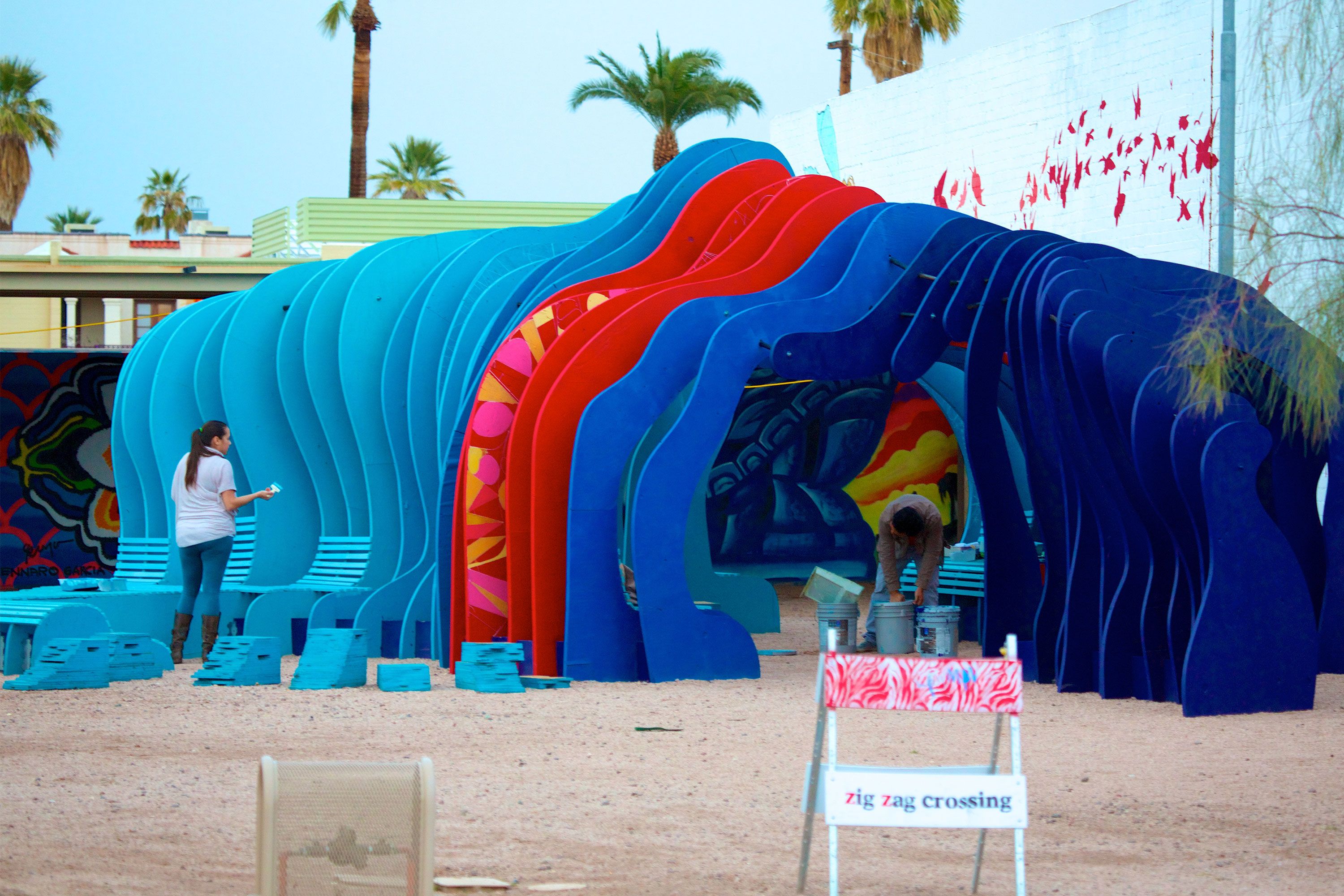 Colorful zig-zag tunnel art installation with people walking and painting in an outdoor space.