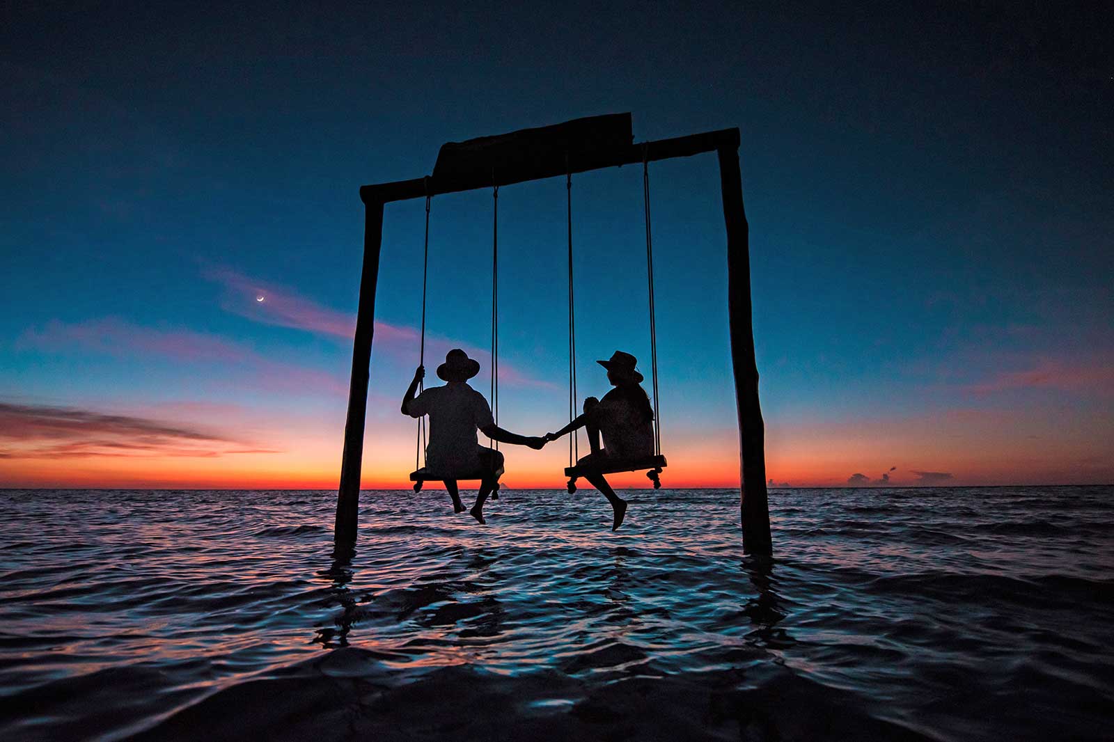 A couple sitting on swings and holding hands at sunset.