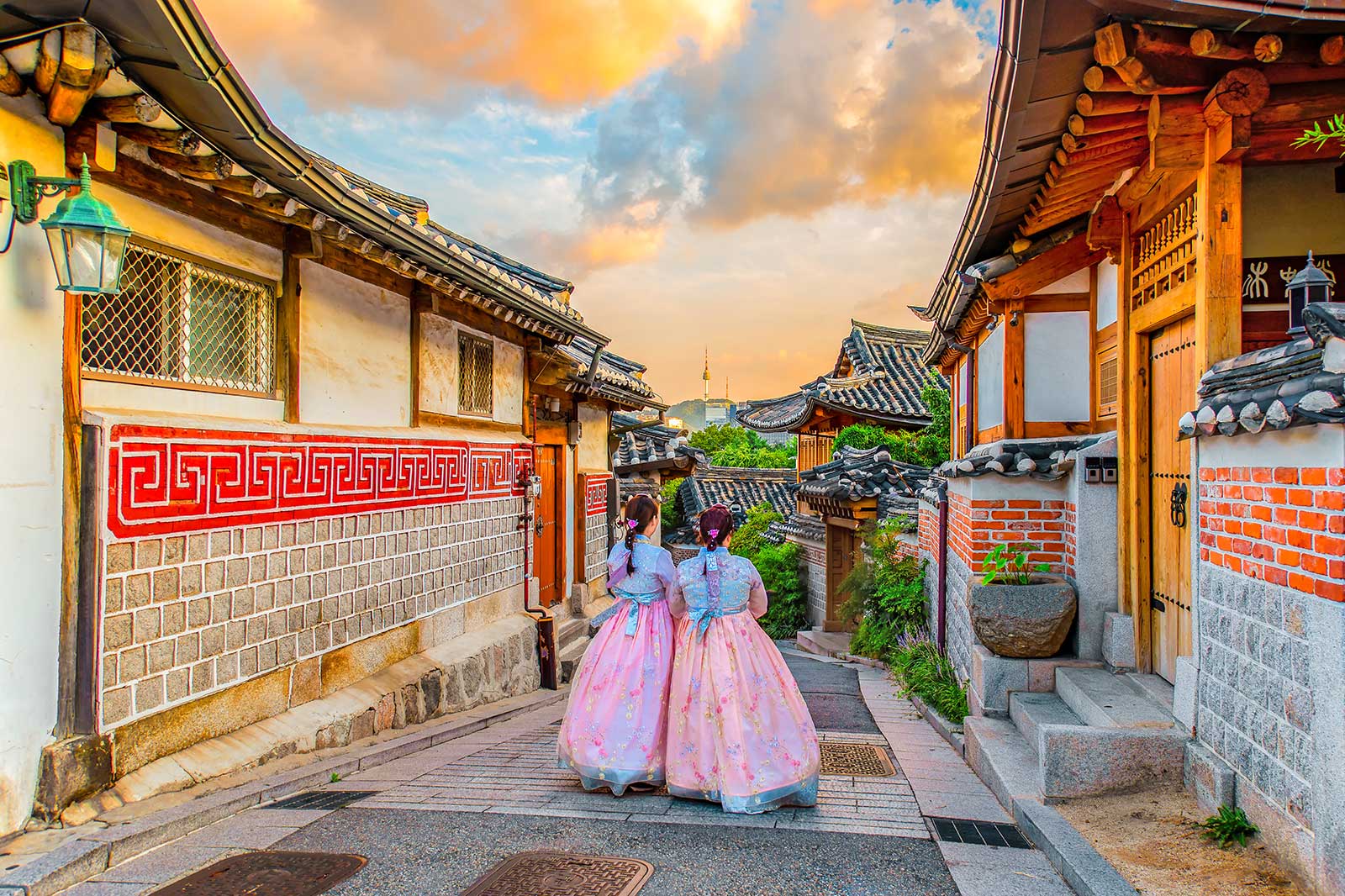 Two people in traditional Korean dresses walking down a narrow lane lined by traditional Korean houses.