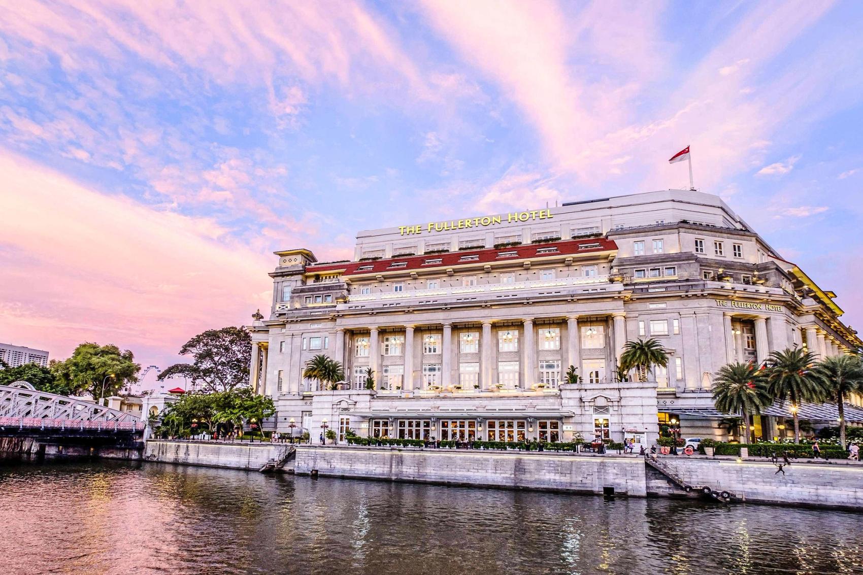The neoclassical Fullerton Hotel in Singapore with water in the foreground and a pink sunset sky in the background.