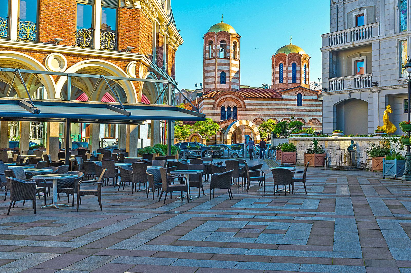 Tables with umbrellas and chair populate a city square with a church in the background.