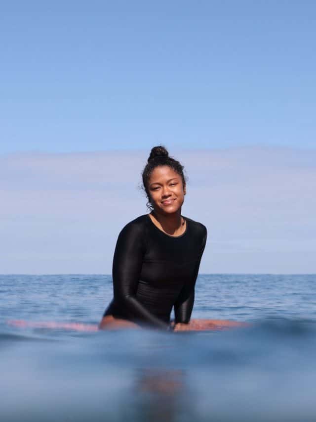 Surfer Chelsea Woody sitting on her surfboard in the sea wearing a black rash vest on a bright, clear day