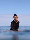Surfer Chelsea Woody sitting on her surfboard in the sea wearing a black rash vest on a bright, clear day