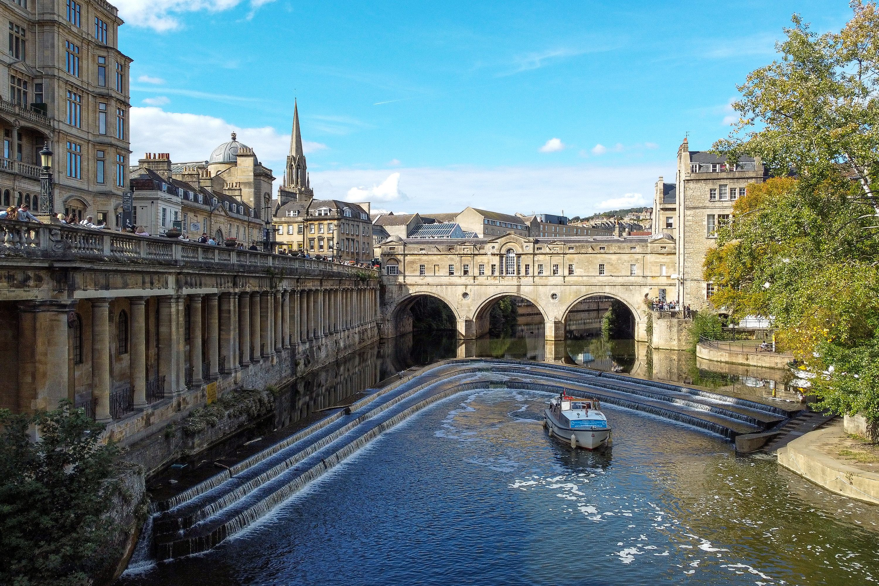 A boat proceeds toward a stone bridge with arched openings and city buildings surround.
