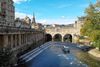 A boat proceeds toward a stone bridge with arched openings and city buildings surround.