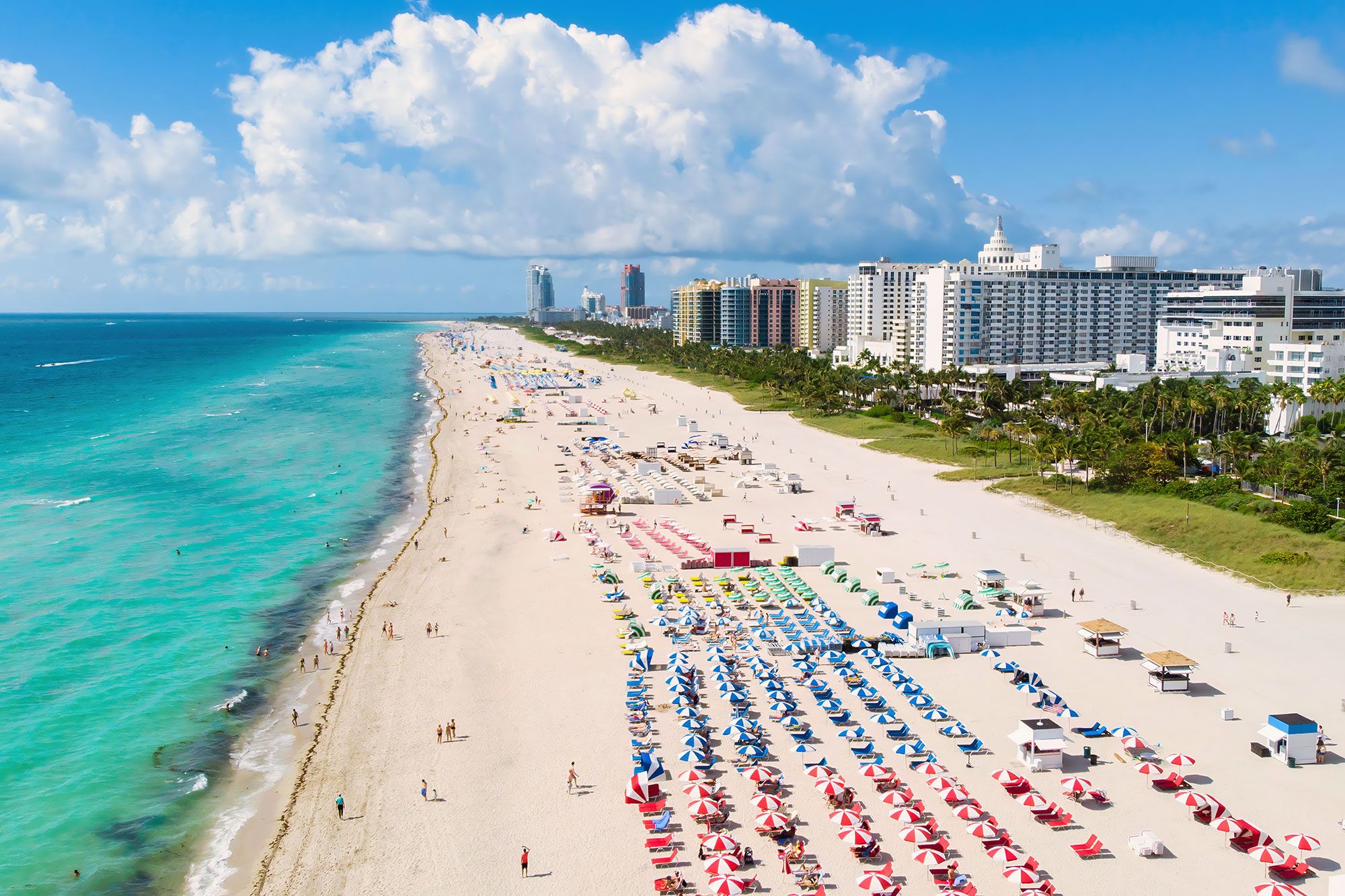 Aerial view of different colored umbrellas on a beach with buildings in the background.