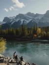 Three people sit on rocks at the edge of the river with fishing equipment as snow-capped Rocky Mountains rise up beyond the opposite bank in Canmore, Canada