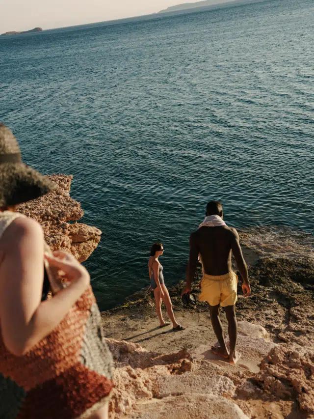 People in swimming gear descend the rocky cliffside down to the sea on a sunny day