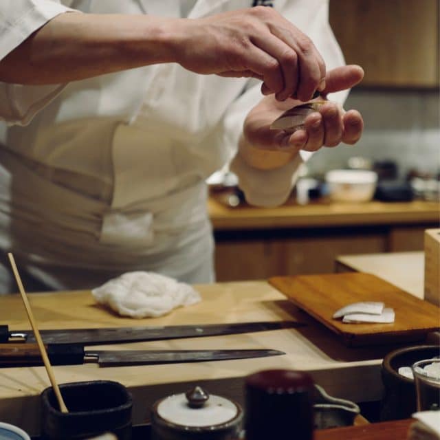 A chef in a white uniform prepares intricate sliced fish and arranges it on a wooden board in a Japanese kitchen