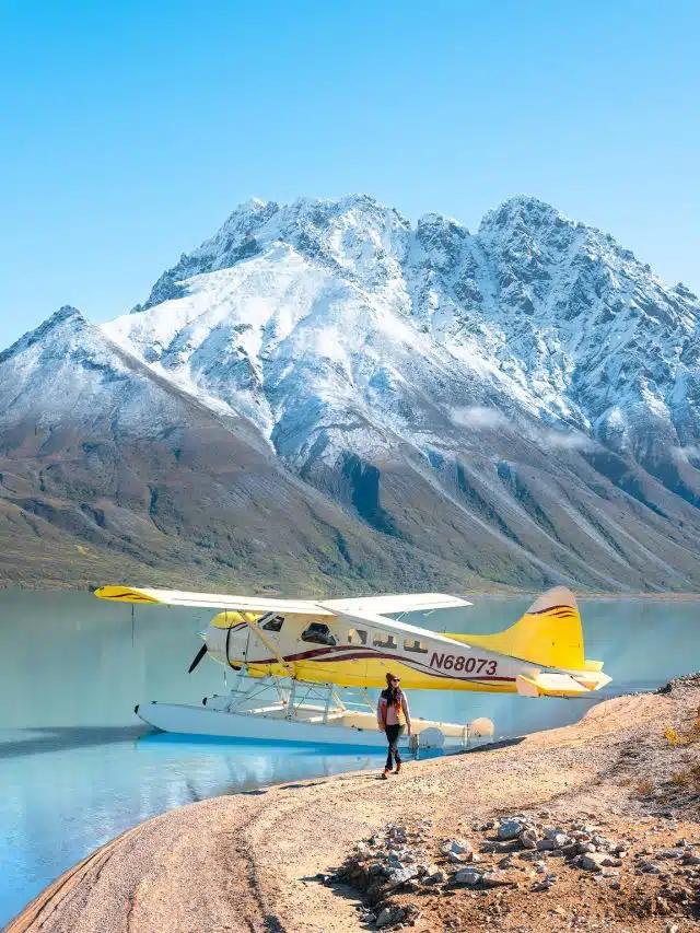 A yellow seaplane is stationary at the edge of the water with a person standing next to it and snow-capped mountains in the background in Lake Clark National Park, Alaska