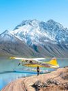 A yellow seaplane is stationary at the edge of the water with a person standing next to it and snow-capped mountains in the background in Lake Clark National Park, Alaska