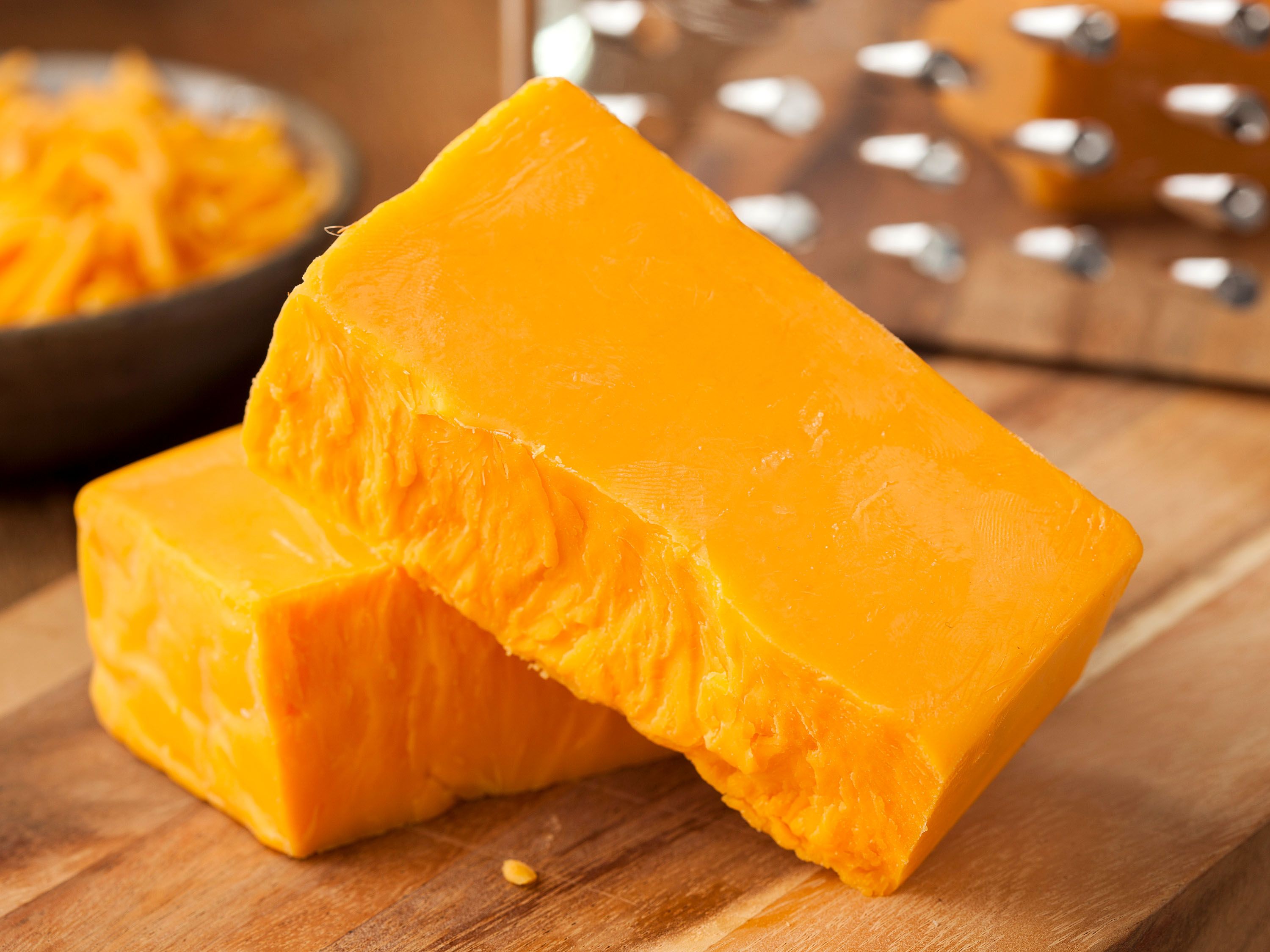 Blocks of cheddar cheese on a wood board with a grater in the background