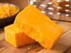 Blocks of cheddar cheese on a wood board with a grater in the background