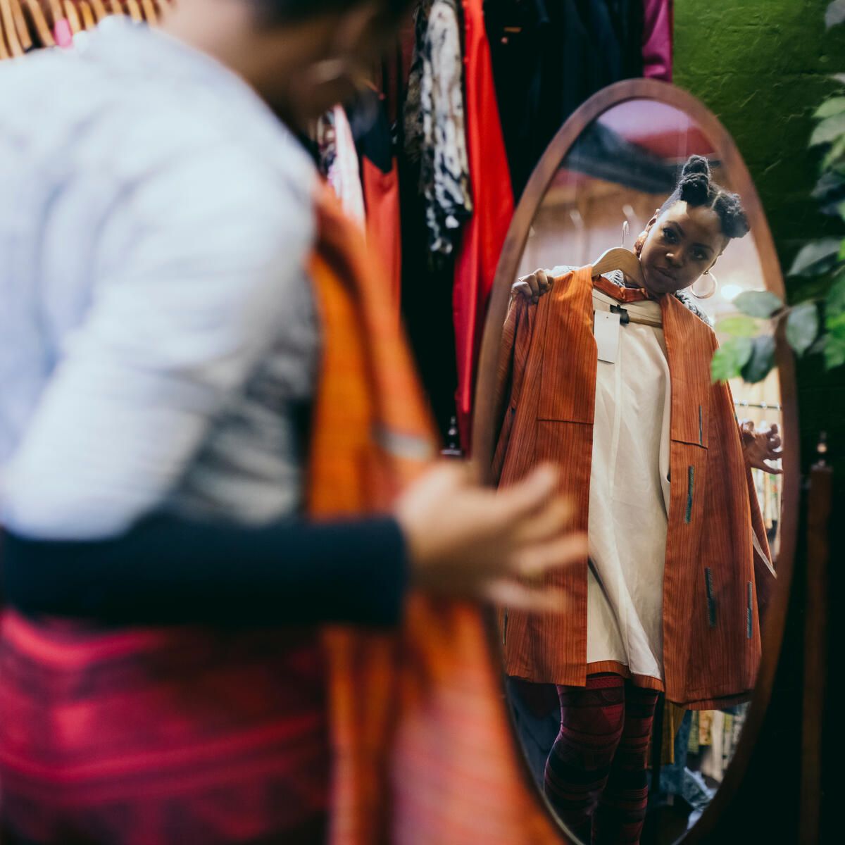 A person holds a dark orange coat up to their body in the reflection of mirror in a vintage clothes store