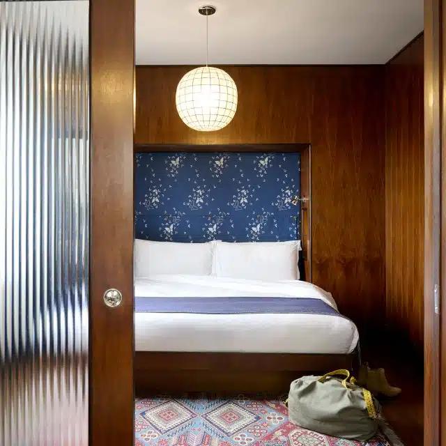 A fluted glass door leads to a hotel room, with wood paneling, a blue patterned headboard, and an overnight bag on the floor