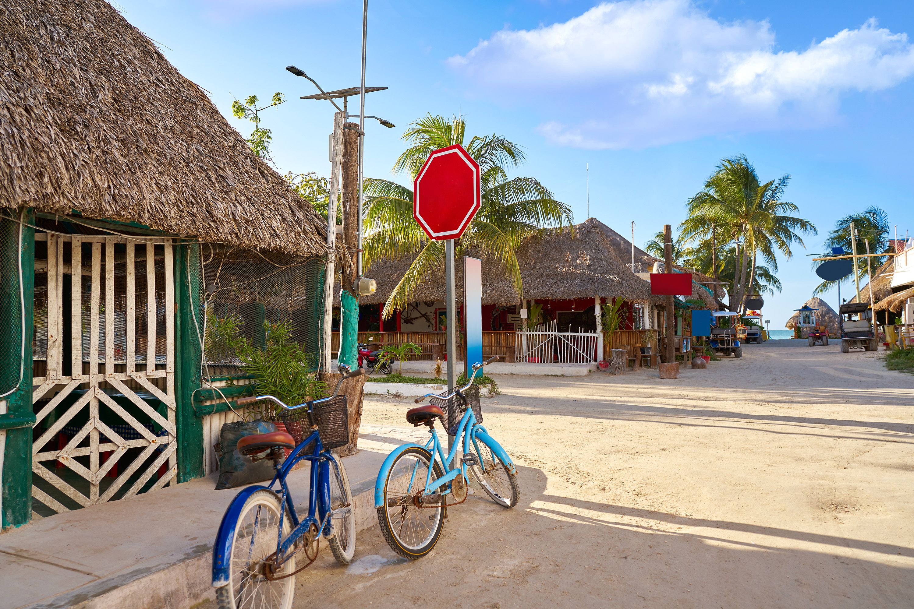 Two bicycles lean against a fence on a sandy street surrounded by buildings with thatch roofs.