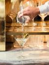 A mixologist's hand pours a martini from a shaker into a glass with two olives on a stick