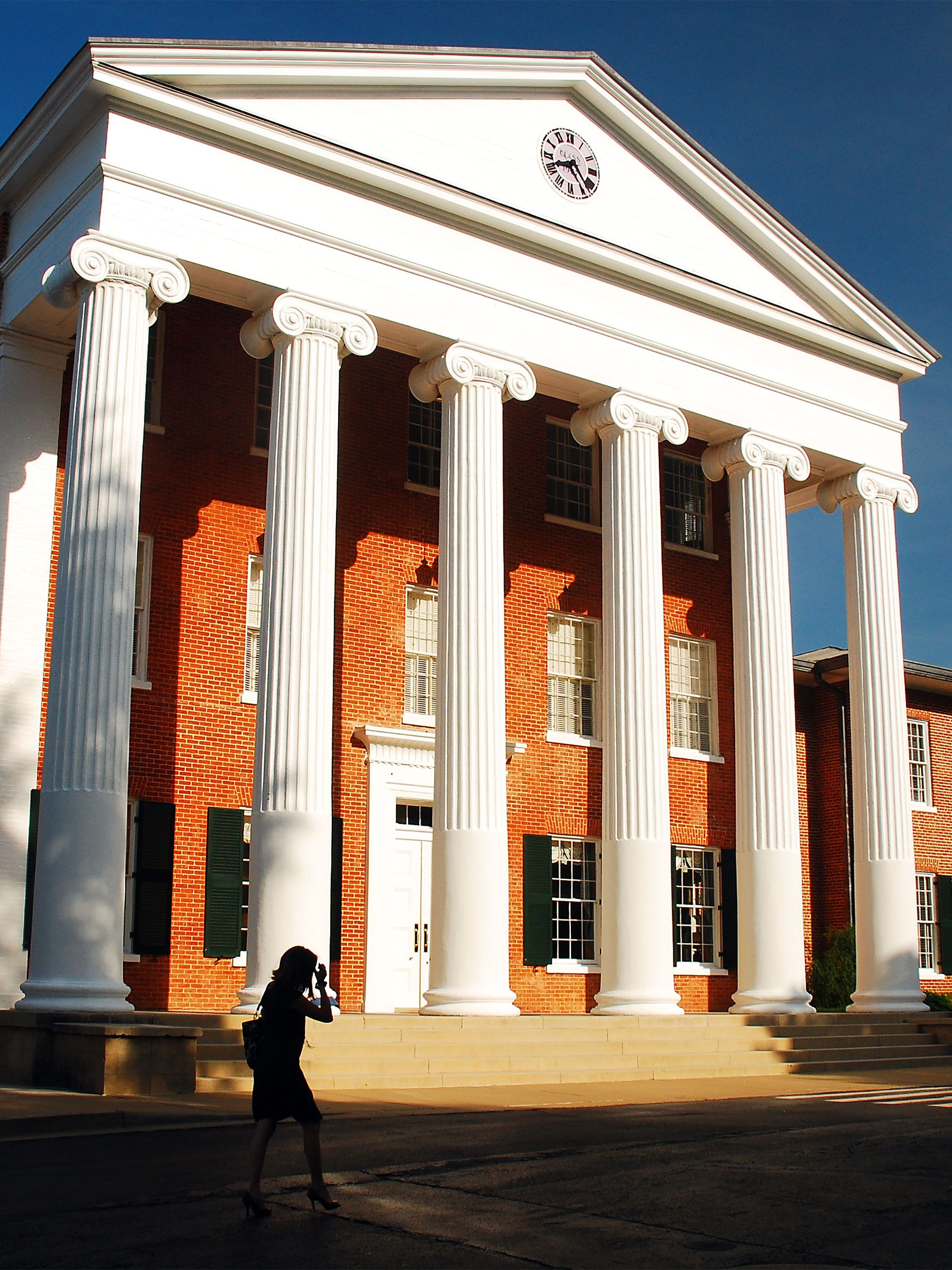 Silhouetted person walks past a red-brick building with tall white columns and clock.