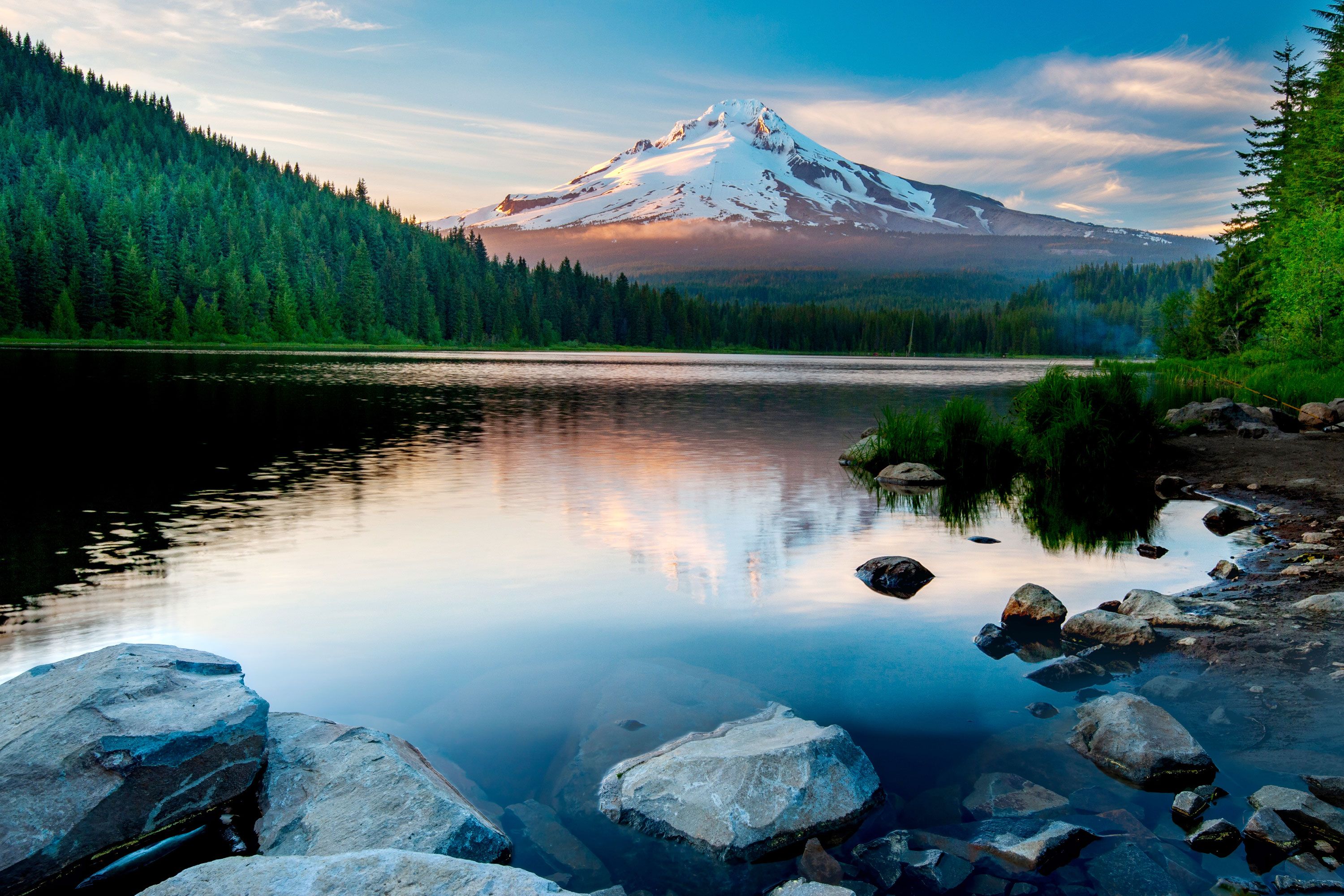 A landscape view of rocks in the foreground of a lake and a snow capped mountain in the distance.