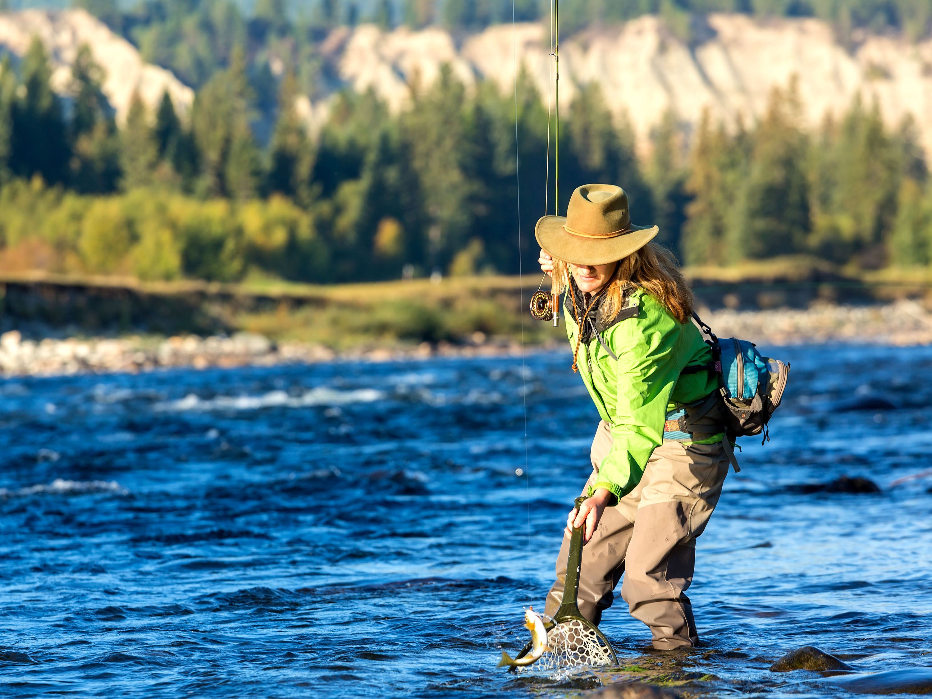 Woman wearing a hat and waders fishing in a stream.