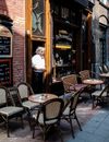 An exterior view of brown café De Pilsener Club in Amsterdam, with a man standing at the doorway and patio tables and chairs on the s