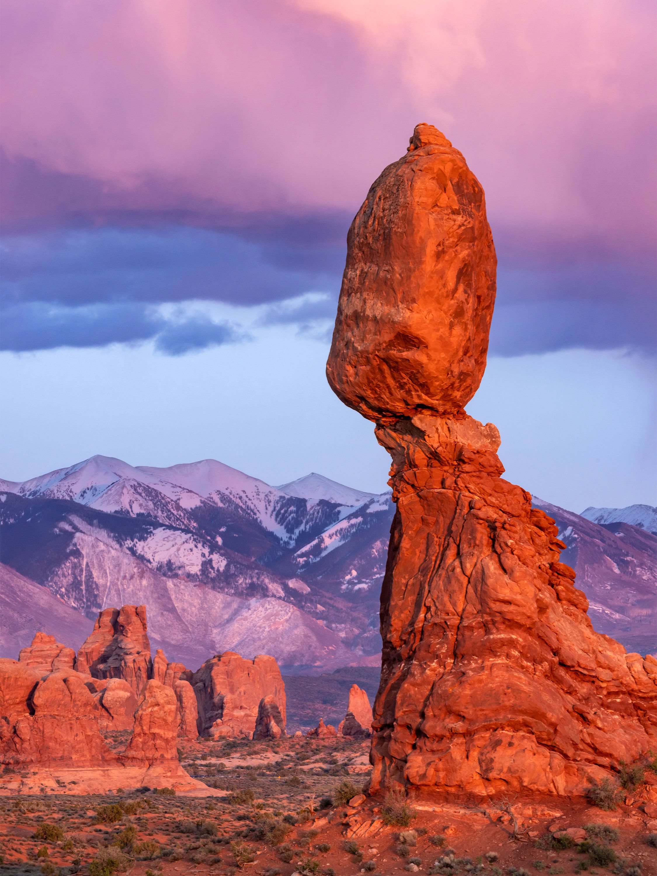 Balanced rock formation glows at sunset with snowy mountains behind.