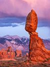 Balanced rock formation glows at sunset with snowy mountains behind.