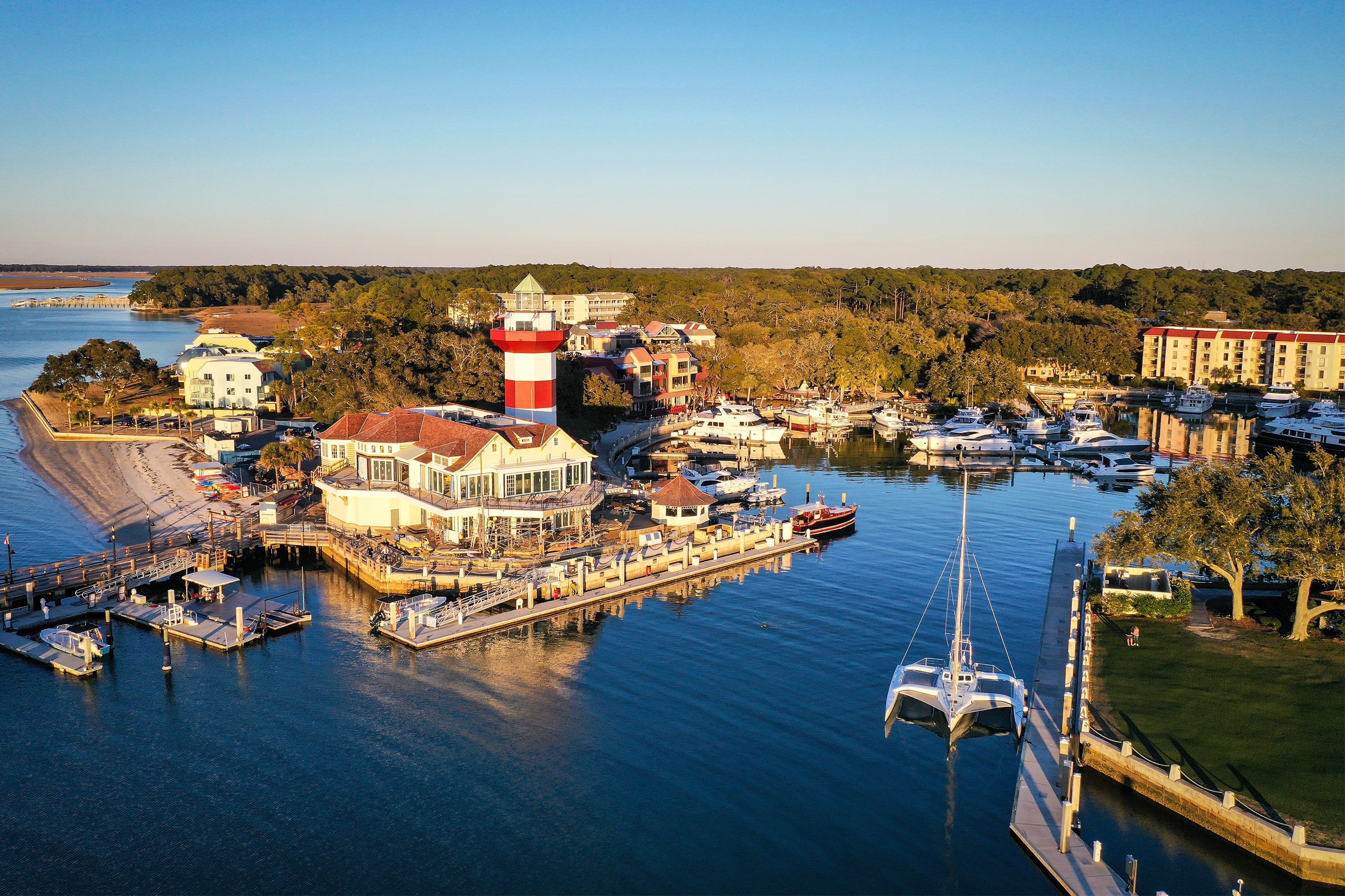 An aerial view of Harbour Town on Hilton Head Island, showing a red-and-white striped lighthouse beside a marina, with surrounding water, docks, boats, and coastal landscape under daylight.
