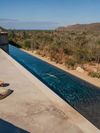 A woman swims in the infinity pool at Paradero Todos Santos, featuring modern architecture, desert landscape, mountains and a hint of ocean in the distance, and a blue sky above