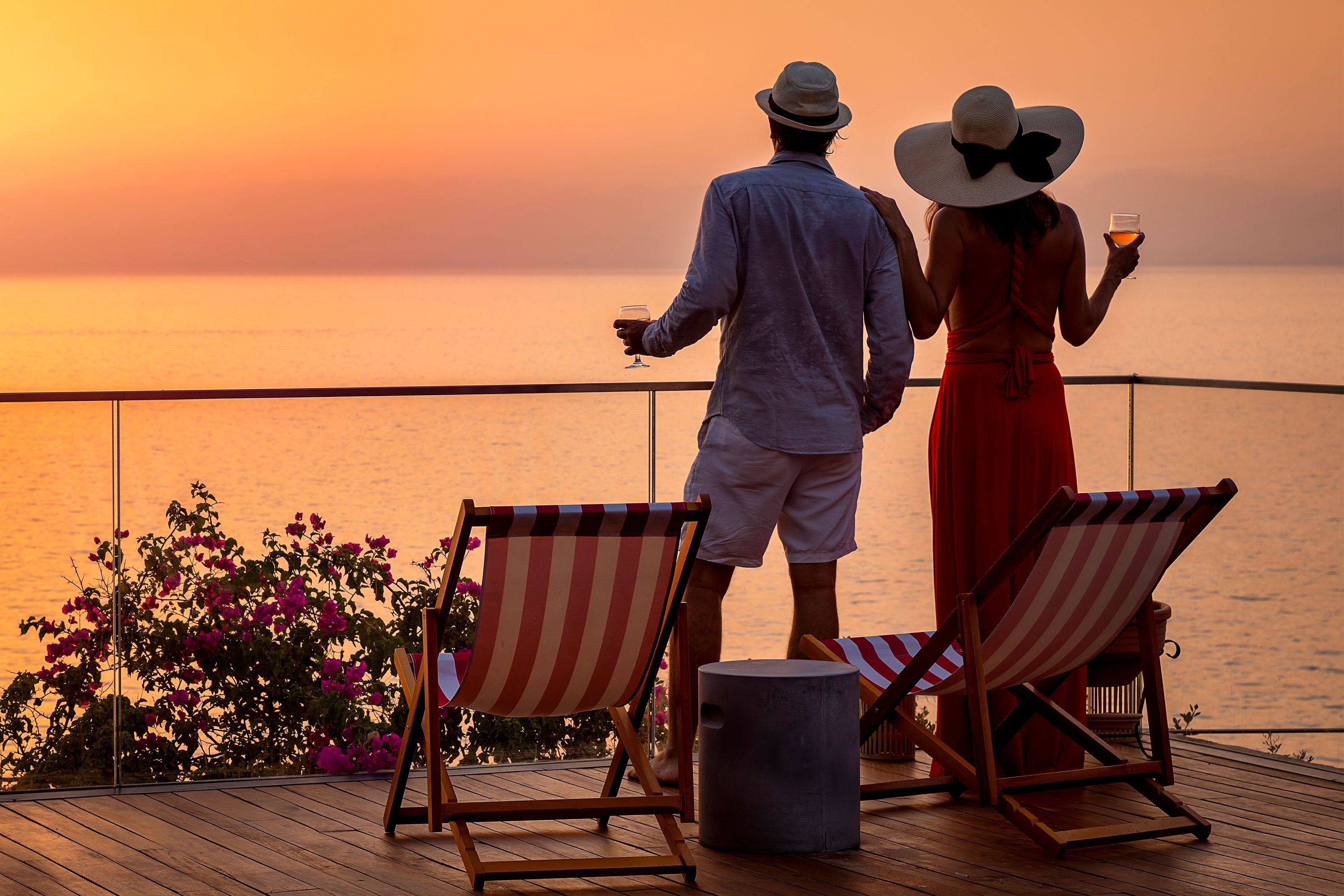 A couple stands on a terrace holding drinks and looking at the ocean at sunset.