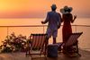 A couple stands on a terrace holding drinks and looking at the ocean at sunset.