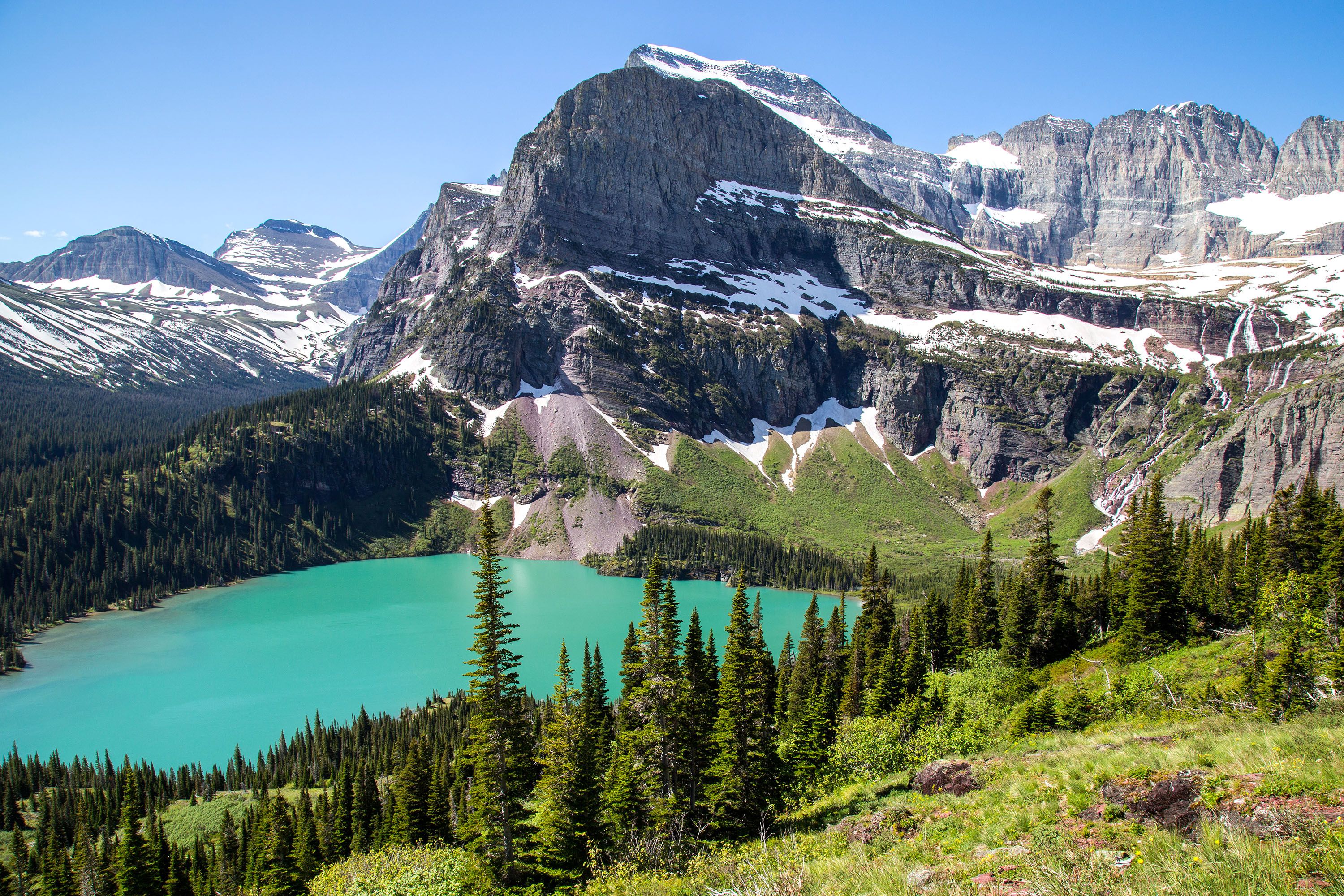 A small turquoise mountain lake, surrounded by fir trees and snow-capped mountains.