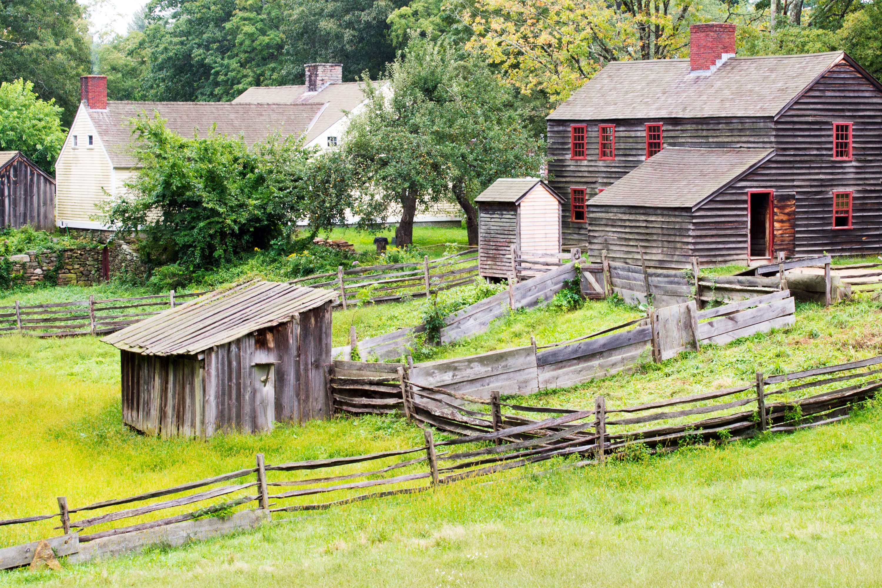 A historic colonial-style farmhouse with adjacent outbuildings surrounded by verdant grass and mature trees under a soft, natural light.