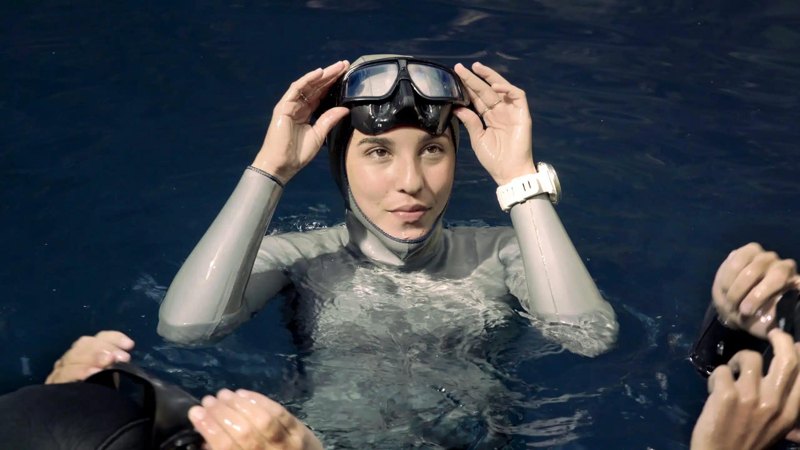 Champion free diver Camila Jaber treads water in a cenote, wearing a grey hooded wetsuit, goggle and white sports watch, with two companions beside her, in Yucatan, Mexico