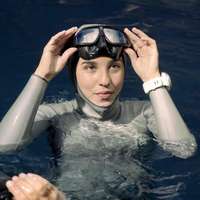 Champion free diver Camila Jaber treads water in a cenote, wearing a grey hooded wetsuit, goggle and white sports watch, with two companions beside her, in Yucatan, Mexico