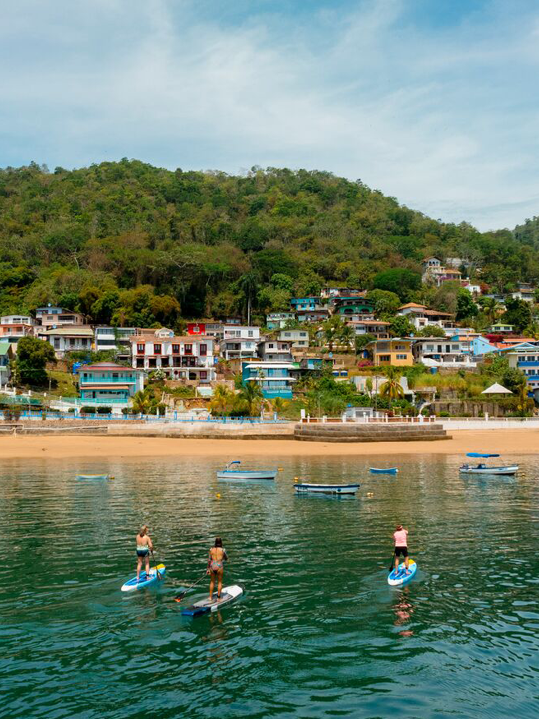 Three stand-up paddleboarders move towards the shore of a golden beach backdropped by a hill