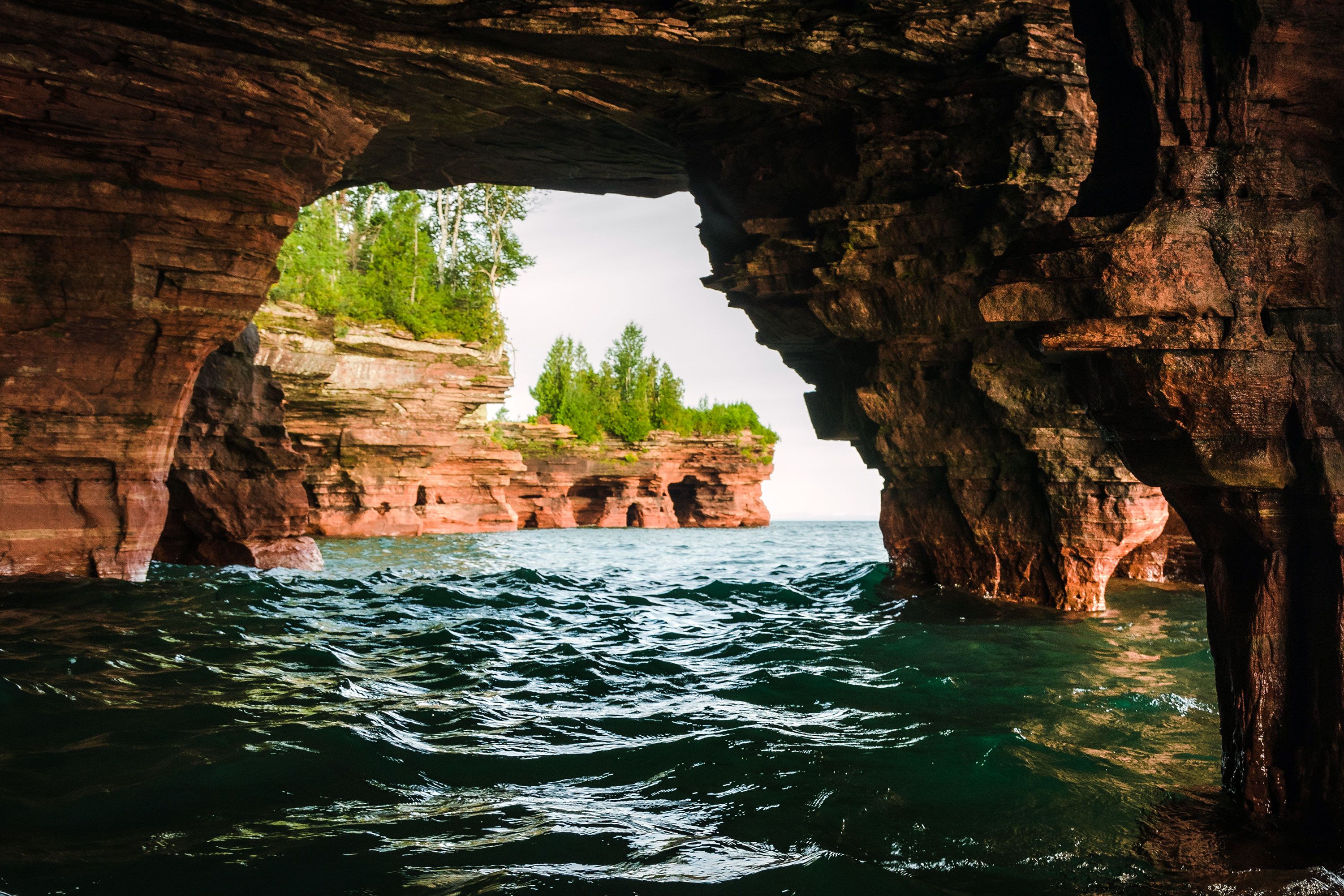 Inside a natural sandstone arch on Devils Island overlooking Lake Superior, reddish brown cave walls frame a view of green water and distant shoreline beneath a partly cloudy sky.