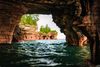 Inside a natural sandstone arch on Devils Island overlooking Lake Superior, reddish brown cave walls frame a view of green water and distant shoreline beneath a partly cloudy sky.