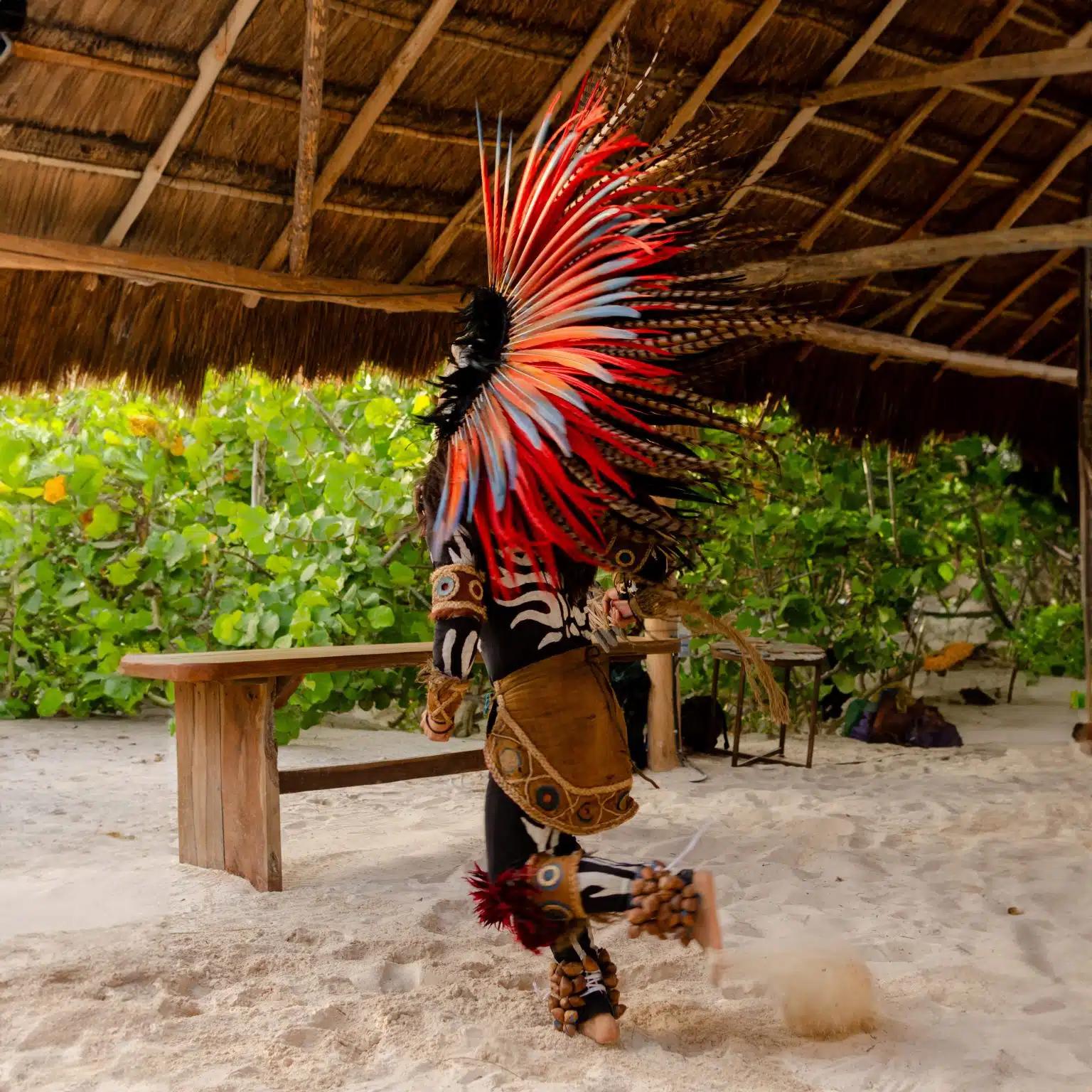A person in a traditional colorful feathered headdress performs on the beach as part of a ceremony in Tulum, Mexico