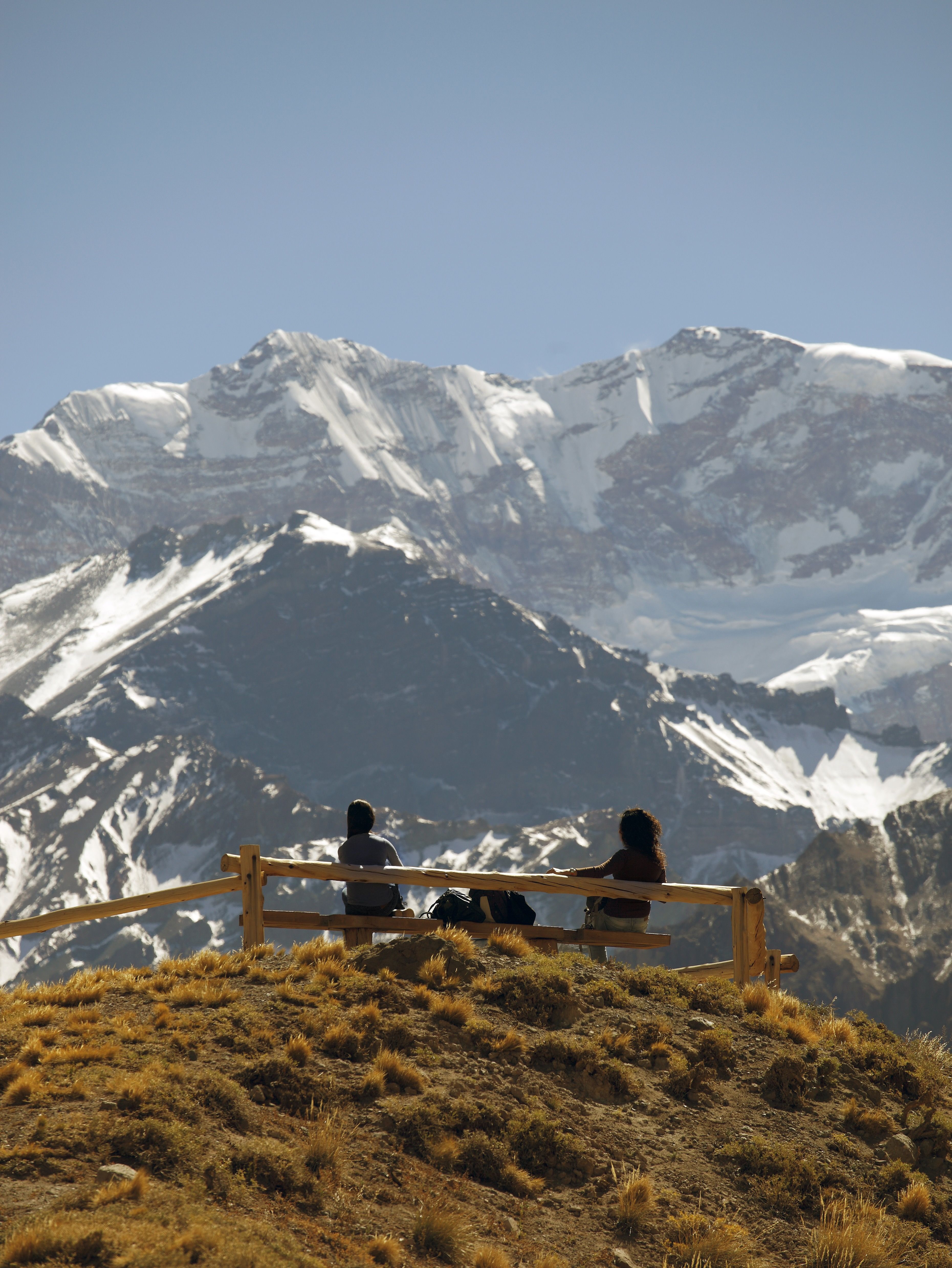 Two people sit on a bench at the top of a grassy bank with a snow-capped Andean mountain in the distance in Mendoza, Argentina